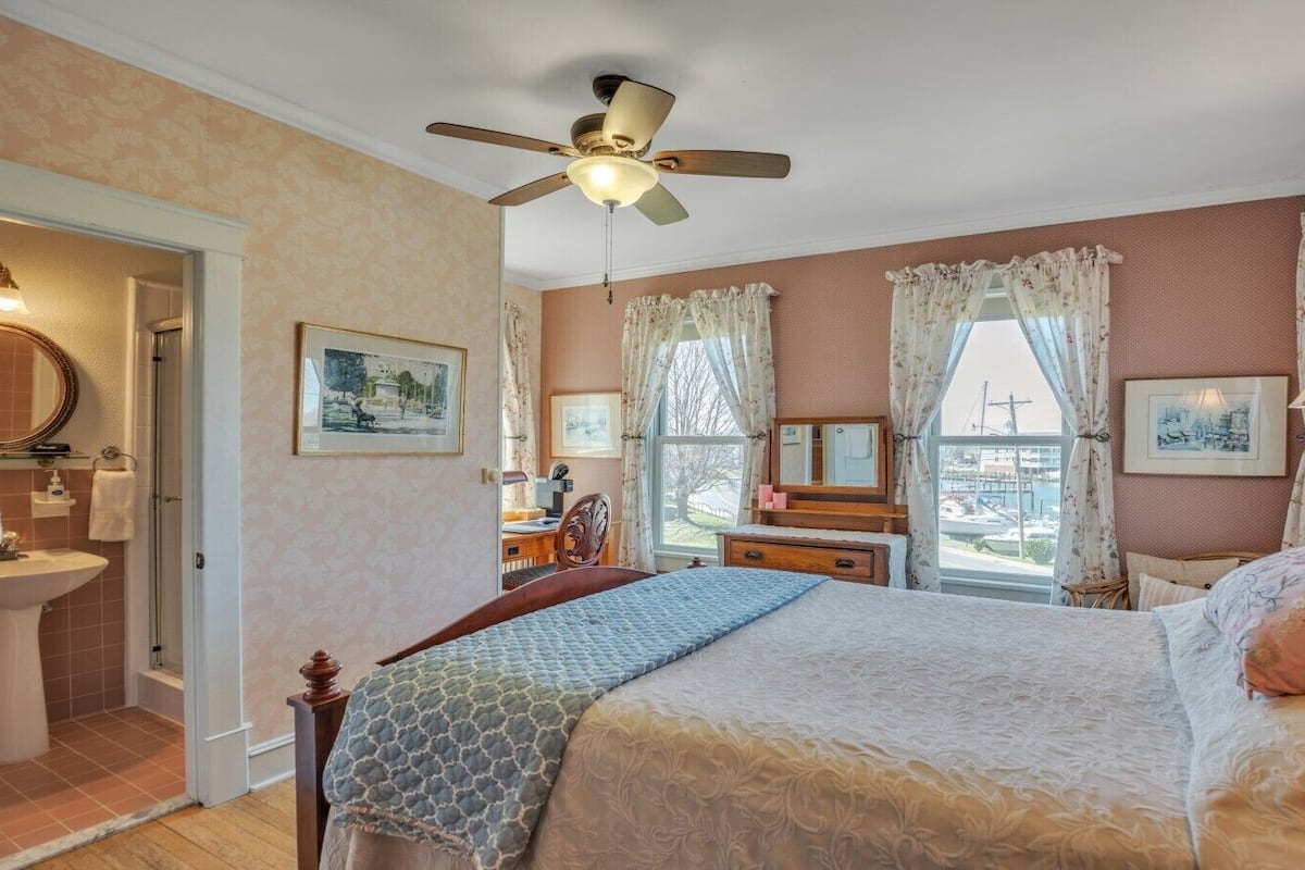 Um quarto com cama, mesa de cabeceira, espelho, janela com cortinas, ventilador de teto e banheiro com pia e chuveiro.