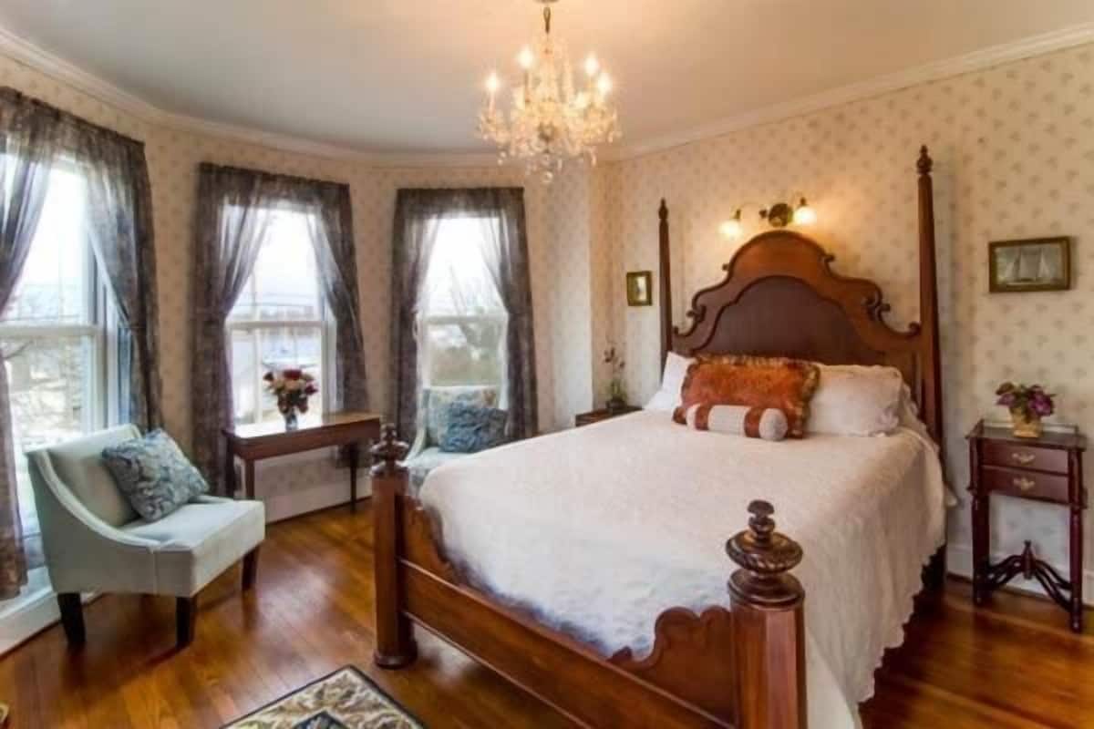 Quarto com cama de madeira baldaquim, lustre e vista para o exterior através de grandes janelas com cortinas.