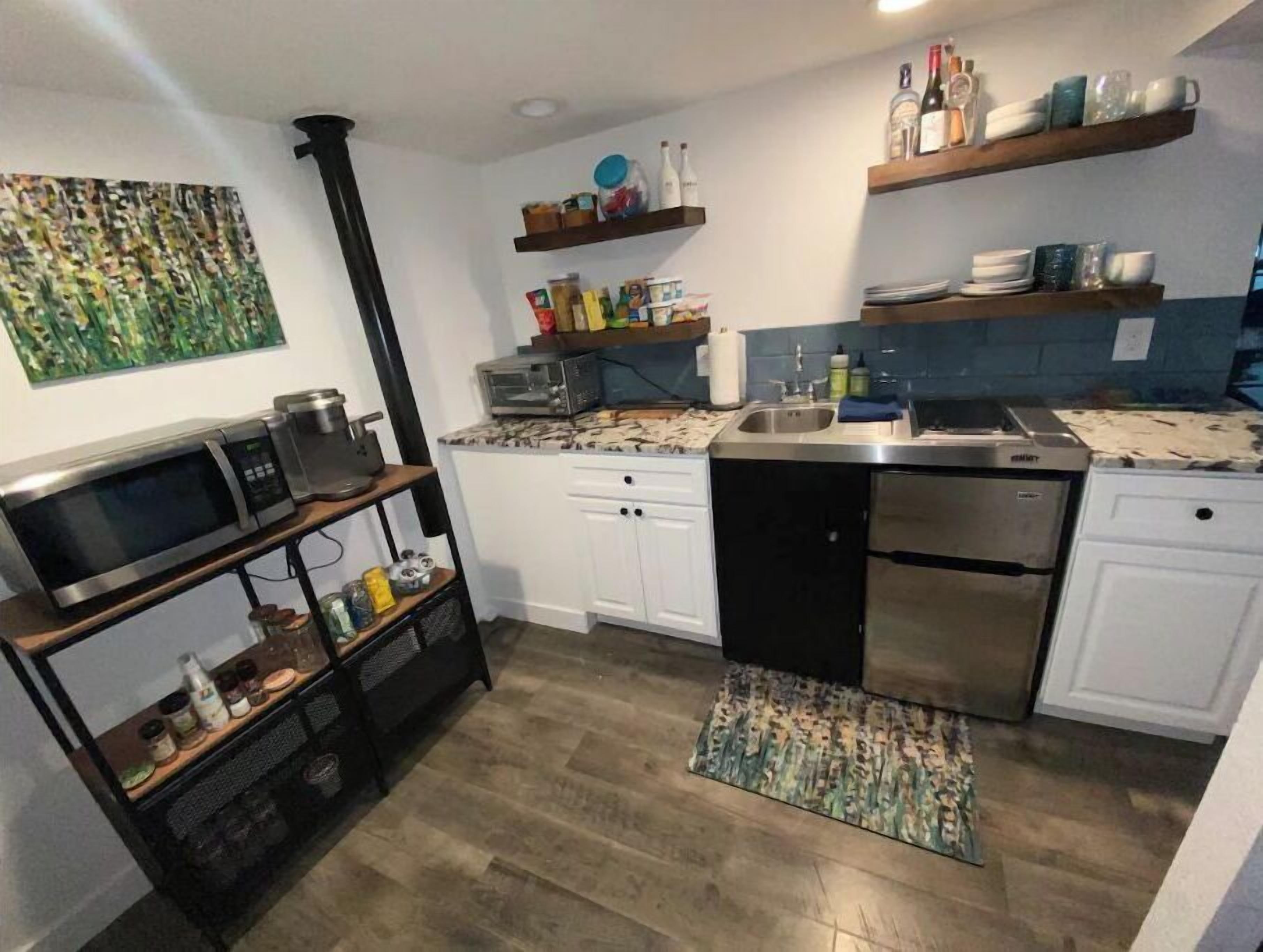 A compact kitchen with white cabinets, a built-in microwave, a dishwasher, and a coffee machine. There are shelves above the counter holding various kitchen items and a small rug on the floor.