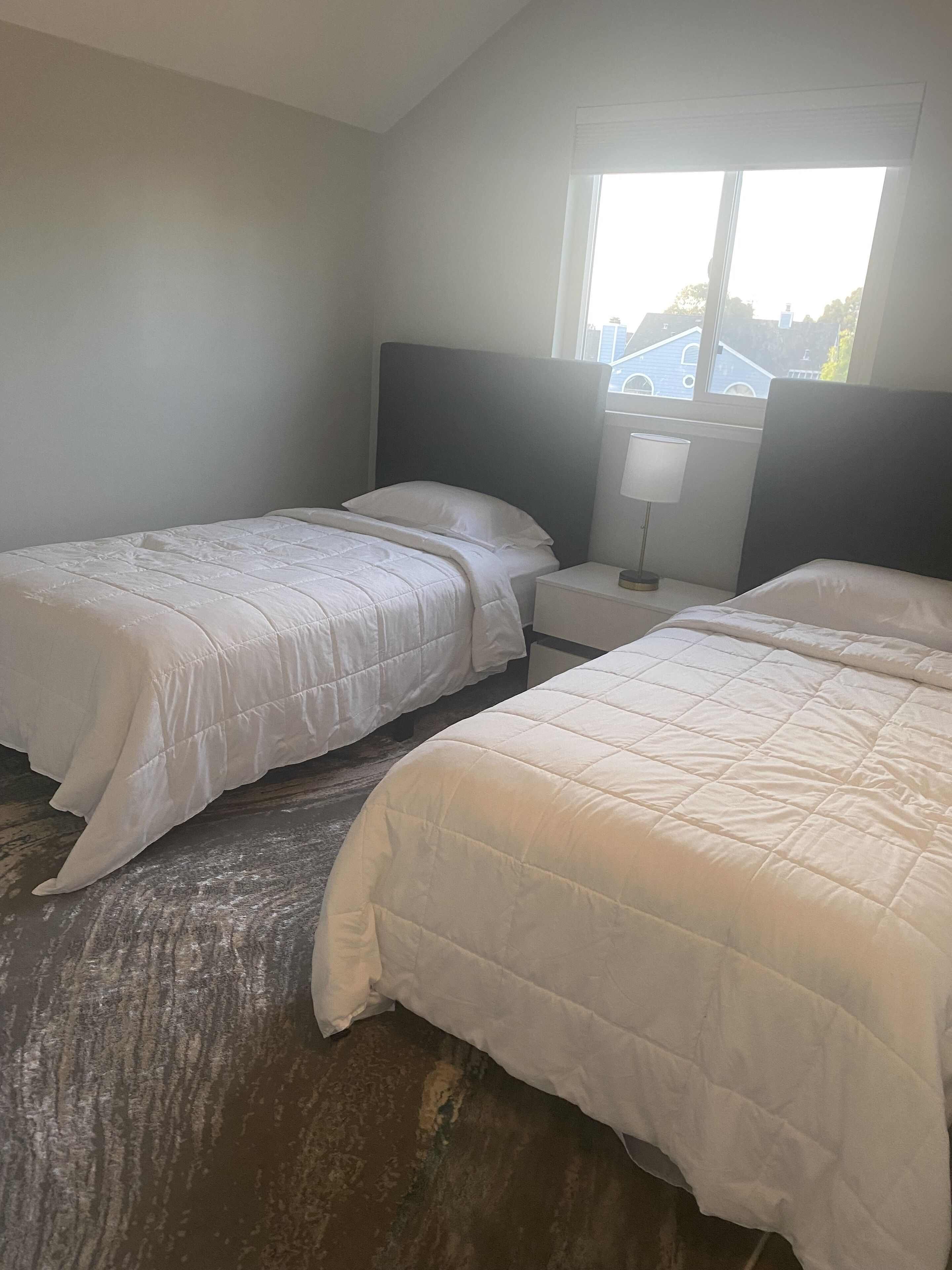 A hotel room with two single beds, a nightstand with a lamp, and a window with a view of buildings.