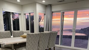 Een eethoek met een tafel en stoelen, grote ramen met uitzicht op een zonsondergang boven het water, en een houten vloer.