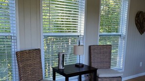 A room with two wicker chairs, a small table with a lamp, and large windows with blinds.