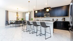 Una cocina moderna con armarios oscuros, encimera blanca y refrigerador de acero inoxidable. Hay cuatro taburetes de bar y una mesa de comedor con sillas.