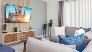 Una sala moderna con una televisión de pantalla plana montada en una pared de madera, un sofá gris con cojines estampados, una mesita auxiliar blanca y un mueble de madera con estantes.