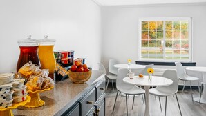 A bright kitchen with white walls, a large window with a view of autumn trees, a buffet counter with food items, and white chairs around a small table. - Hotel One75 (Hamilton)