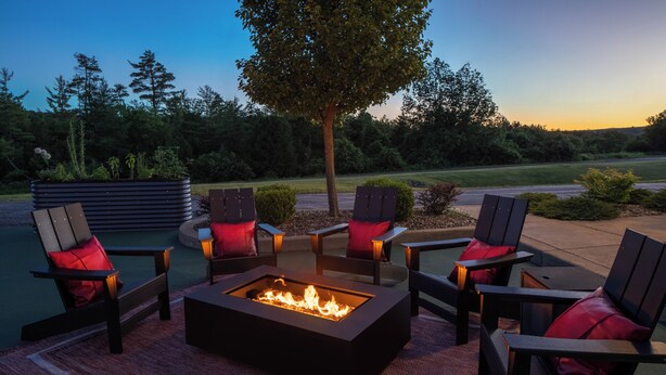 A fire pit surrounded by four Adirondack chairs with red cushions, set on a patterned rug. - Hotel One75 (Hamilton)