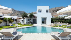 Un área de piscina moderna con sillones y sombrillas, rodeada por un edificio blanco y un paisaje montañoso.