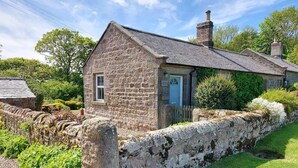 A stone cottage with a small porch and a chimney, surrounded by a stone wall and greenery. - Cosy, Tranquil, Fully Equipped & Beautiful Garden (Chathill)