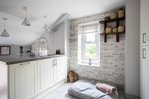 Una cocina con armarios blancos, pared de ladrillo, ventana y una cama tipo altillo con una toalla rosa.