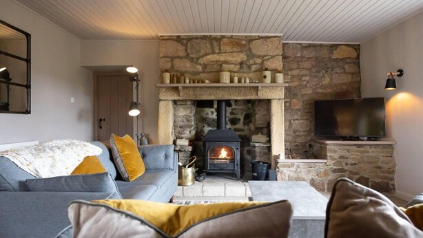 A cozy living room with a stone fireplace, a grey sofa with yellow cushions, and a flat-screen TV on a stone wall. - Cosy, Tranquil, Fully Equipped & Beautiful Garden (Chathill)