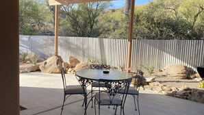 A covered patio area with a round metal table and four chairs, surrounded by a concrete floor and a corrugated metal fence.