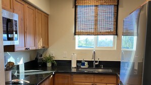 A kitchen with wooden cabinets, a black countertop, a stainless steel refrigerator, and a window with blinds.