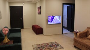 A living room with a sofa, a red ottoman, a TV mounted on the wall, and a door leading to another room. - Magic Suite Mahboula-5 (Mahboula)