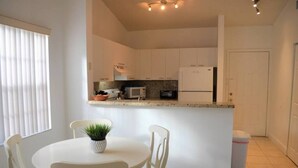 Una cucina compatta con pensili bianchi, un forno a microonde e un frigorifero. C'è un piccolo angolo ristoro con tavolo e sedie, e un vaso con una pianta sul tavolo.
