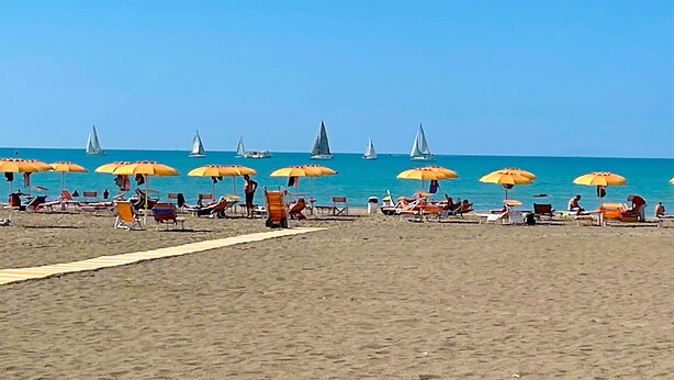 Ein Strand mit Reihen von Sonnenschirmen und Liegestühlen, einem Sandweg und Segelbooten auf dem Wasser.