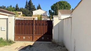 Une entrée fermée par un portail mène à une propriété avec une allée en gravier, un mur blanc et, au fond, un bâtiment dont le toit est recouvert de tuiles.