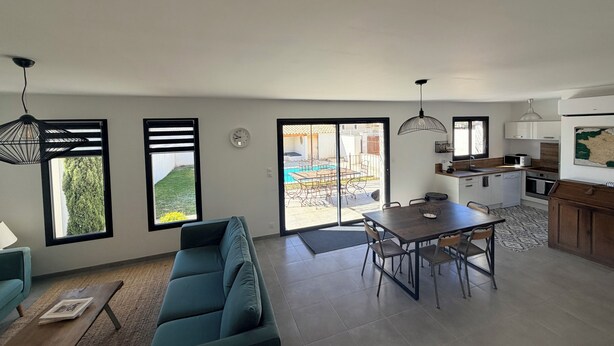 Un salon moderne avec un canapé, une table à manger et des chaises, une cuisine équipée de meubles et d’appareils électroménagers, et une vue sur une piscine à travers une porte vitrée.
