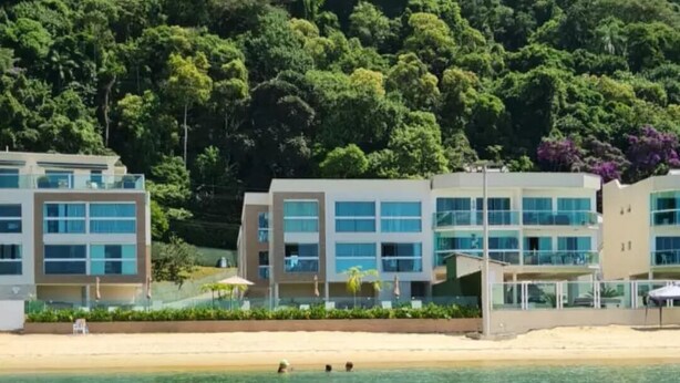 Un hotel frente al mar con varias construcciones, una playa arenosa y una colina boscosa al fondo.