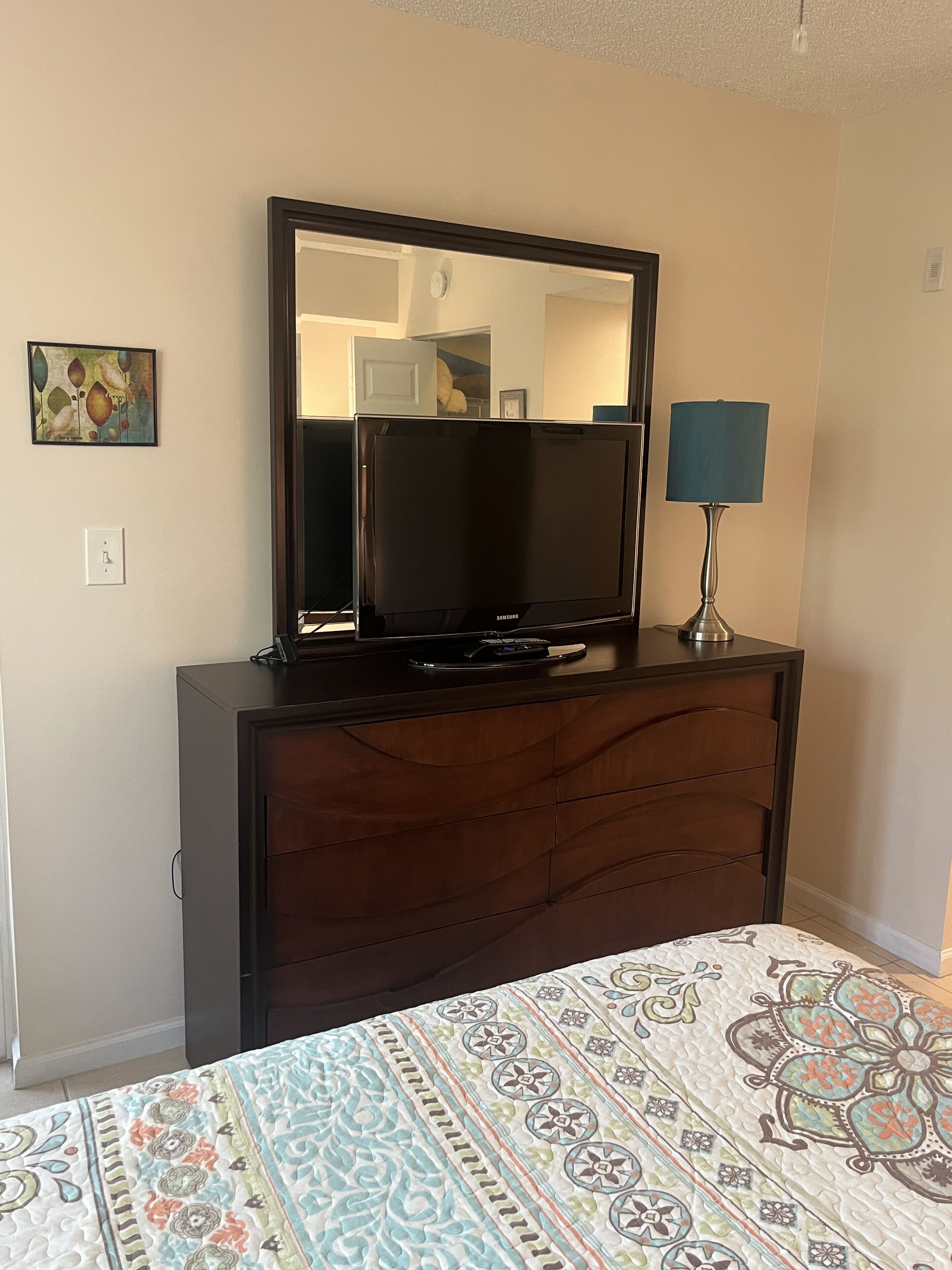 A bedroom with a bed, a dresser with a television, a lamp, and a painting on the wall.