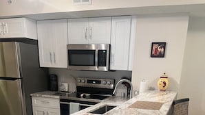 Une cuisine moderne avec des meubles blancs, un réfrigérateur en acier inoxydable, un four à micro-ondes et une cuisinière. On y trouve deux tabourets de bar en osier et une petite table sur laquelle est posé un vase.