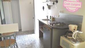 A compact kitchen with a gas stove, microwave, and a small sink. A chair is placed near the window, and a door is visible in the background. - 1 Bedroom Apartment in Aubenas (Aubenas)