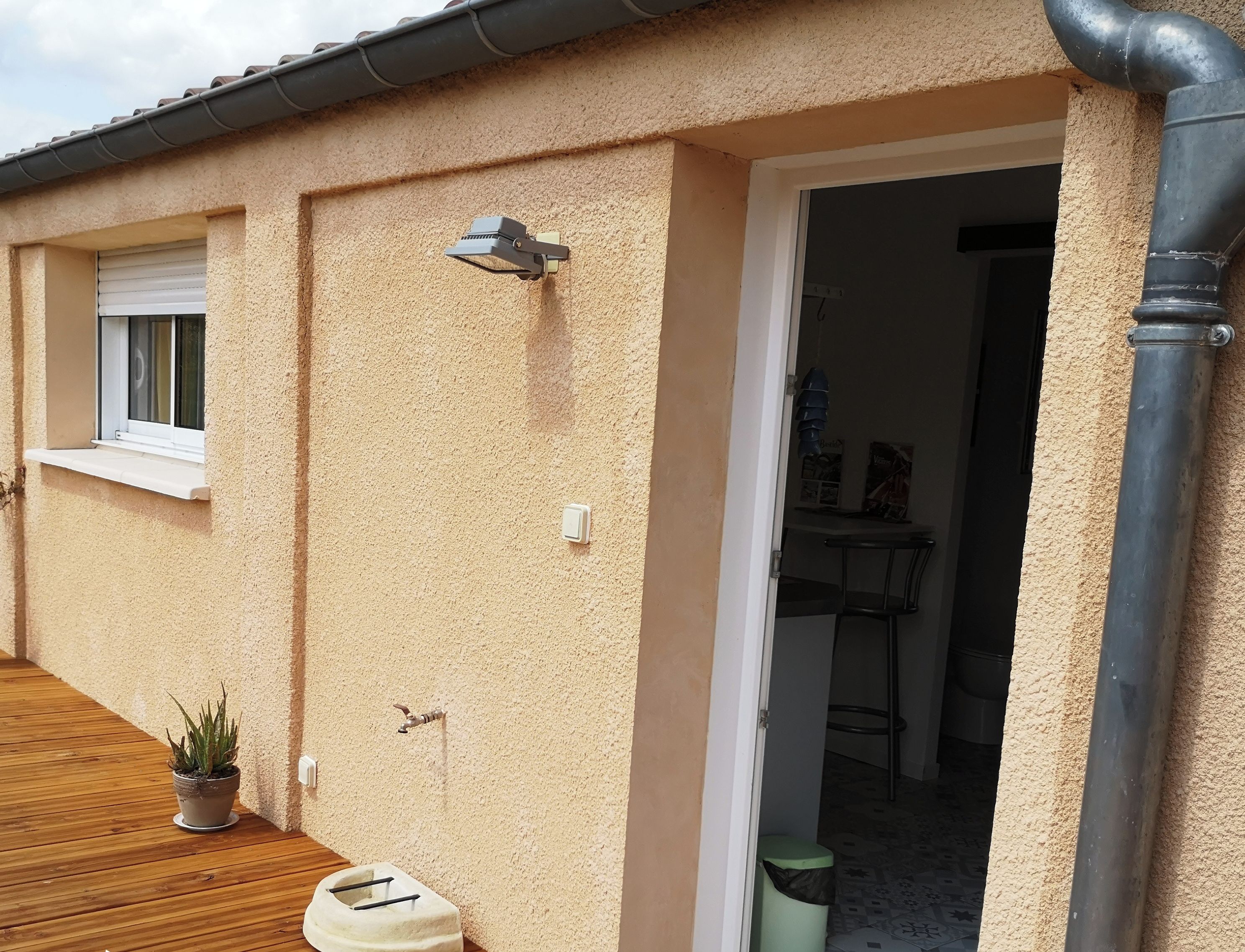 L’extérieur d’un bâtiment avec une terrasse en bois, une fenêtre, un luminaire fixé au mur et une porte donnant sur l’intérieur.