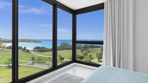 Habitación con amplias ventanas que dan a una playa y al océano, cama con cubierta azul claro y cortina blanca a la derecha.