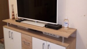 En platt-TV står på ett TV-bord i trä, tillsammans med en fjärrkontroll och en flaska handsprit. Under TV-bordet finns ett vitt skåp med lådor.