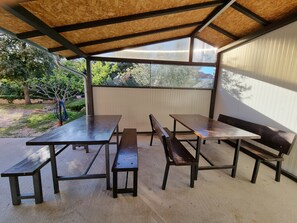 Een terras buiten met metalen tafels en banken, een overdekt gedeelte met een houten plafond, en een uitzicht op bomen en een gebouw op de achtergrond.