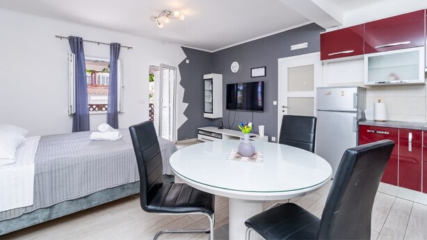Una habitación de hotel moderna con cama, una mesa redonda de comedor con sillas, televisión y una kitchenette con armarios rojos.