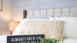 Un lit bien fait, agrémenté d’oreillers à motifs et d’une pancarte sur laquelle on peut lire : « DIEU TOUT-PUISSANT, DIEU D’AMOUR, RÉJOUIS-TOI EN CET ENDROIT ».