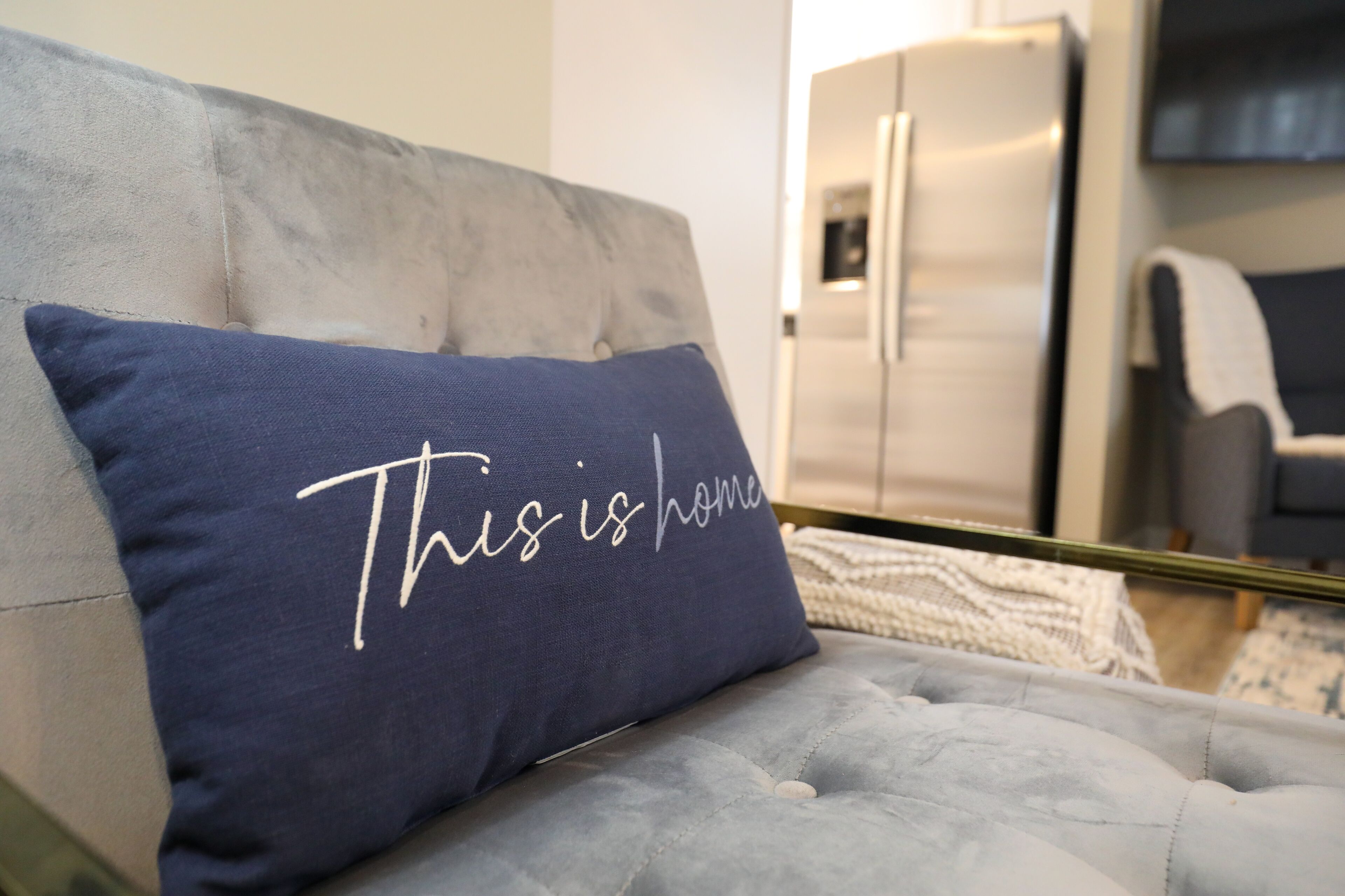 A tufted sofa with a navy blue pillow that reads "This is home."