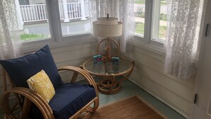 Una habitación de esquina con una silla de mimbre, una mesita con lámpara y vistas al exterior.