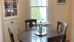 Un comedor con una mesa redonda, cuatro sillas, un armario con puertas de cristal, una ventana con persianas y un cuadro en la pared.