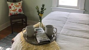 Una cama bien hecha con edredón blanco, una bandeja con dos tazas blancas y un periódico, una pequeña planta en maceta y un cuadro en la pared.