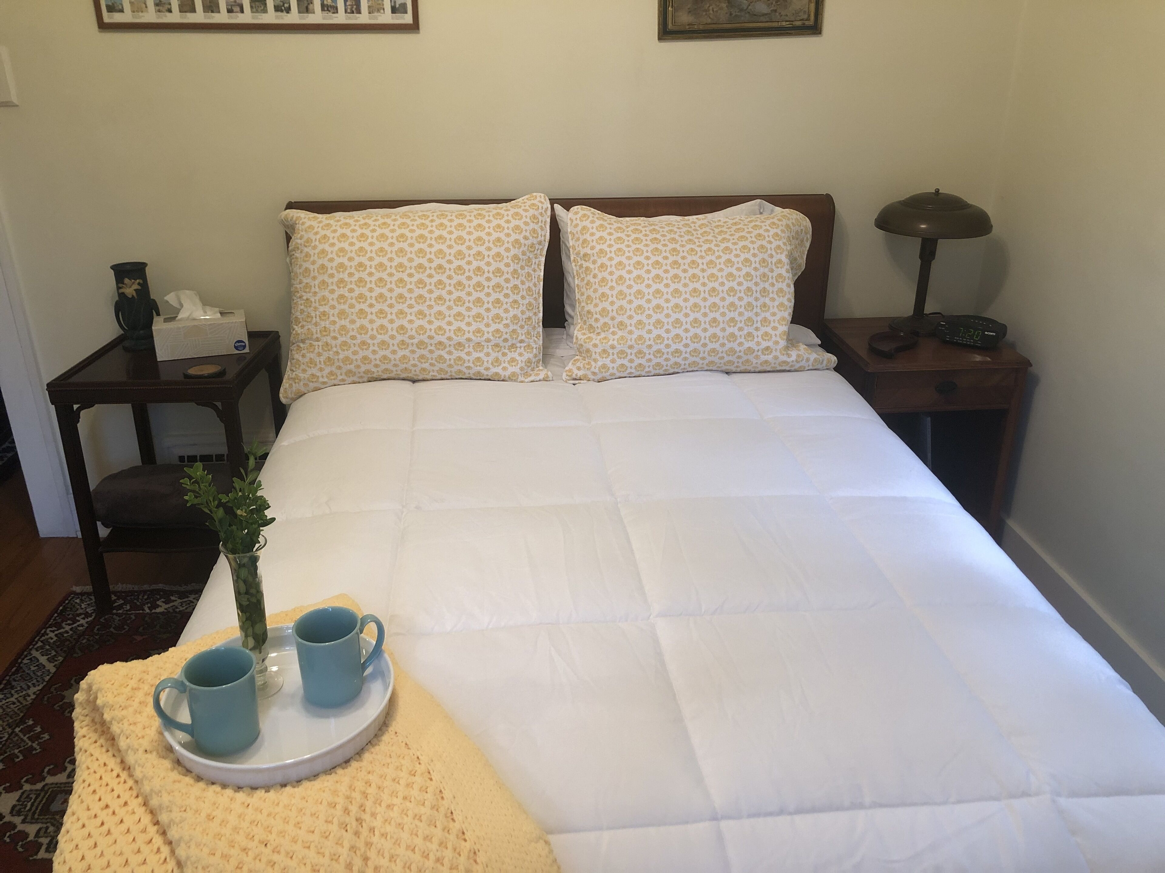Un lit bien fait, avec du linge de lit blanc et des coussins à motifs, une table de chevet avec une lampe et une horloge, une petite plante et deux tasses bleues sur un plateau.