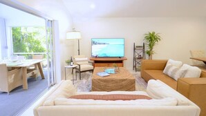 A modern living room with a large TV, comfortable seating, a dining area, and a view through large windows. - 594 Sandy Shore Château w Heated Pool | Walk2Beach (Naples)