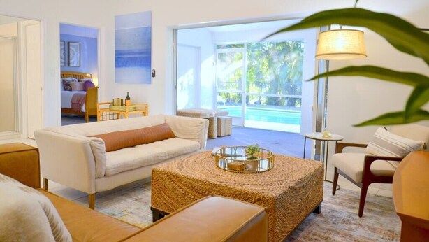 A modern living room with a sofa, a coffee table, and a lamp. There is a view of a pool through large windows. - 594 Sandy Shore Château w Heated Pool | Walk2Beach (Naples)
