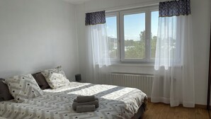 A bedroom with a bed, two pillows, a bedside table, a lamp, and a window with curtains. - Sélestat Beautiful house with large wooded garden Meublé tourisme Les Tulipes 3⭐ (Sélestat)