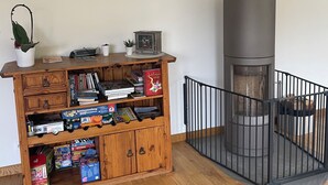 A room with a wooden bookshelf filled with books and games, a small plant, and a modern fireplace. - Sélestat Beautiful house with large wooded garden Meublé tourisme Les Tulipes 3⭐ (Sélestat)
