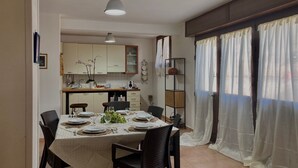 Et spiseområde med et bord dækket til fire personer, et køkken i baggrunden og et vindue med lette gardiner.