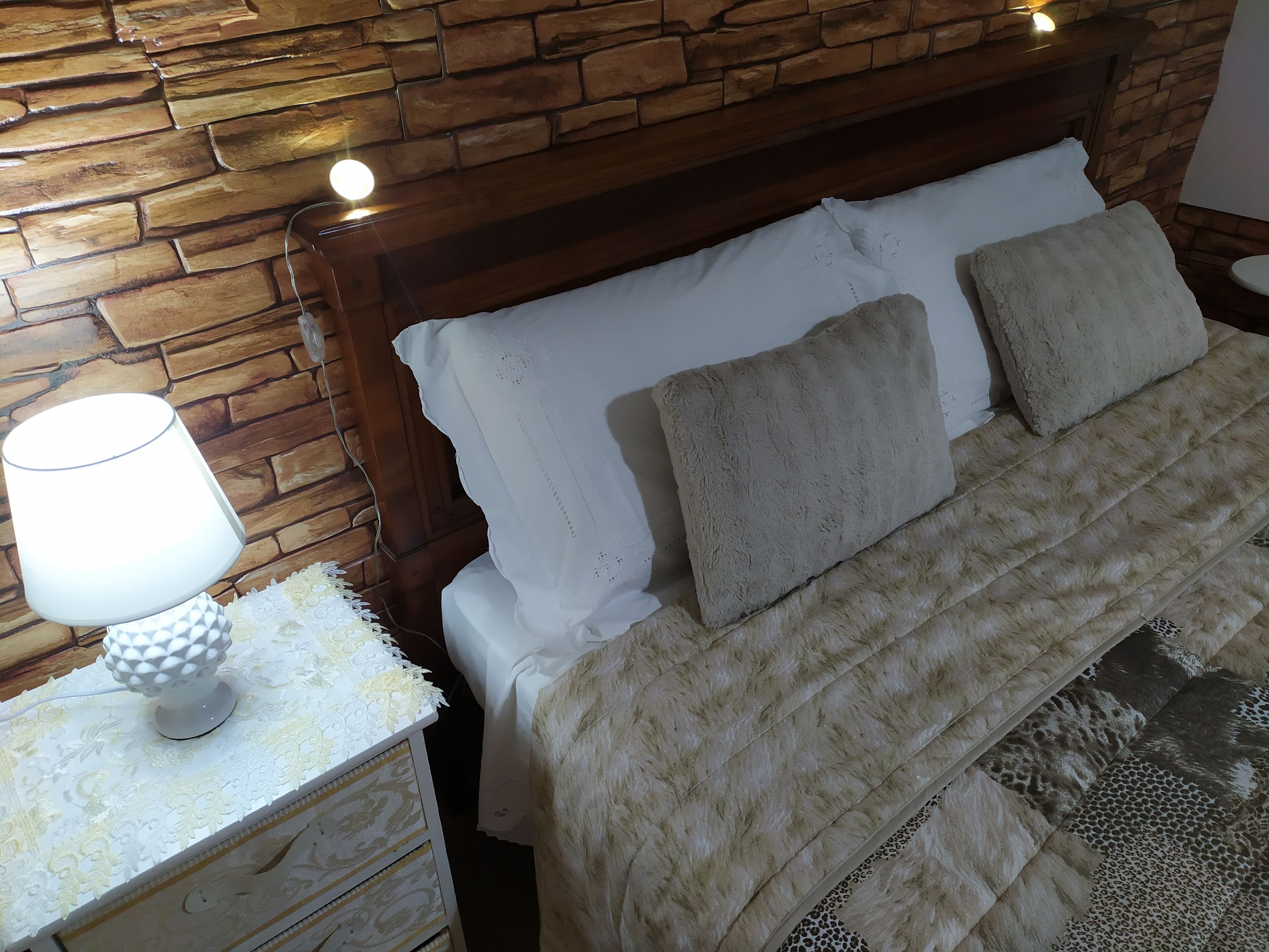 A bedroom with a stone wall, a wooden headboard, a white bed with fluffy pillows, a nightstand with a lamp, and a decorative table.