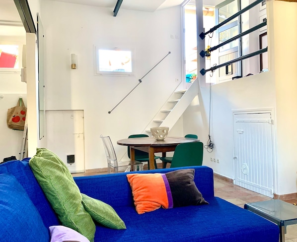 Una sala de estar con un sofá azul, una mesa de comedor con sillas, una escalera y un televisor montado en la pared.
