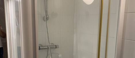 A shower enclosure with a glass door, a handheld showerhead, a wall-mounted faucet, and a small shelf with toiletries.