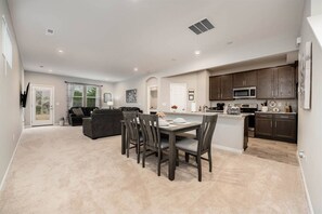 A spacious living area with a dining table and chairs, a kitchen with dark cabinets and appliances, and a door leading to a balcony.