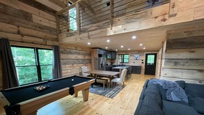 Interior de una cabaña de madera con mesa de billar, sofá, comedor y cocina.