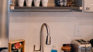 Cucina con zoccolo schienale in piastrelle bianche, elettrodomestici in acciaio inossidabile e mensole con tazze, bicchieri e una pianta.
