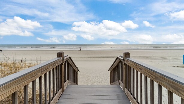 Una escalera de madera que conduce a una playa, con cielo despejado y algunas personas a lo lejos.