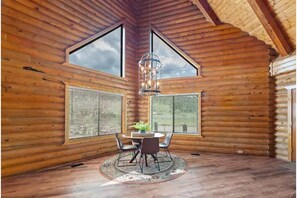 Interior de una cabaña de madera con comedor que incluye una mesa, sillas, ventanas amplias y una lámpara colgante.