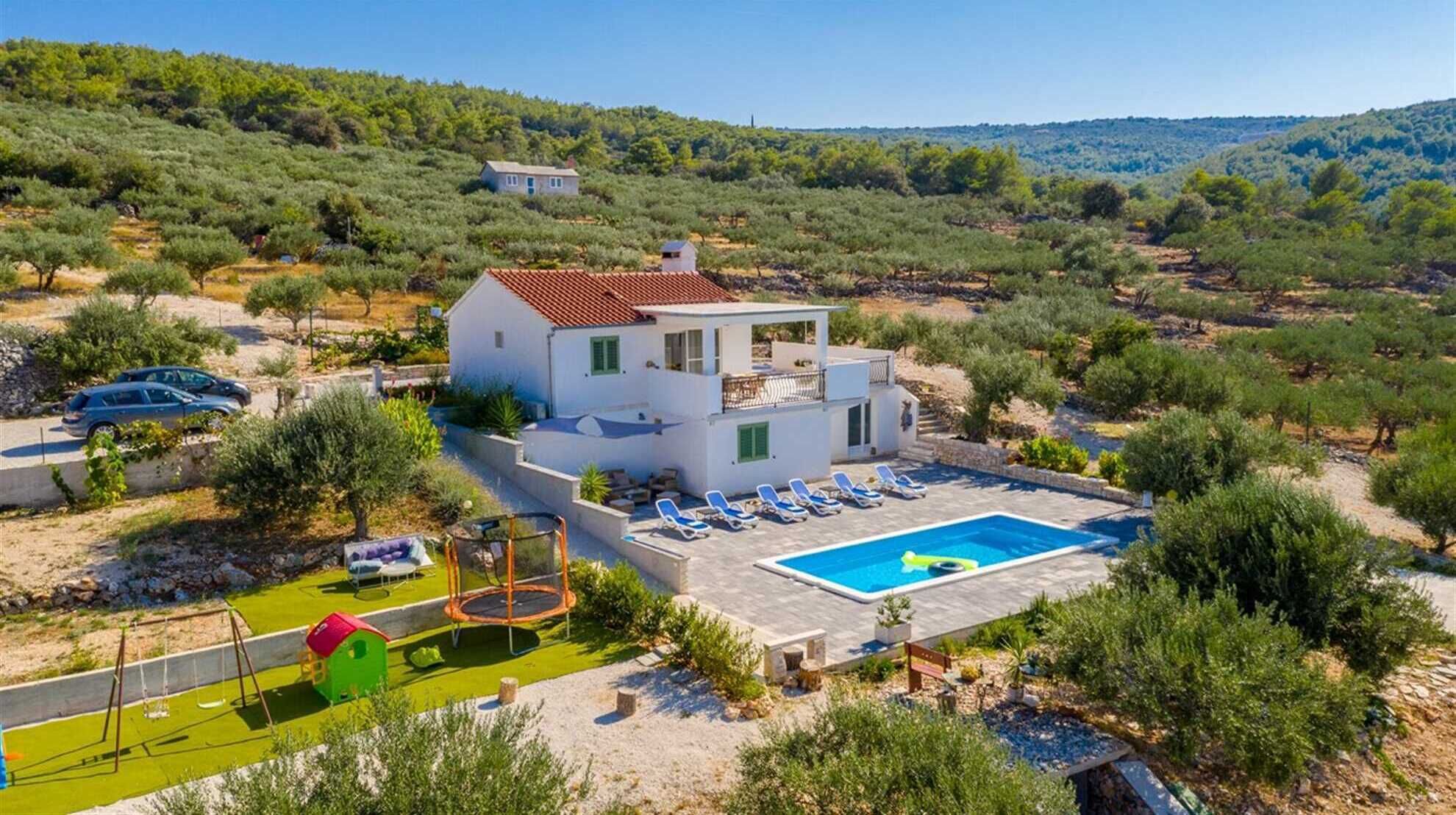 A two-story house with a swimming pool, surrounded by greenery and hills.